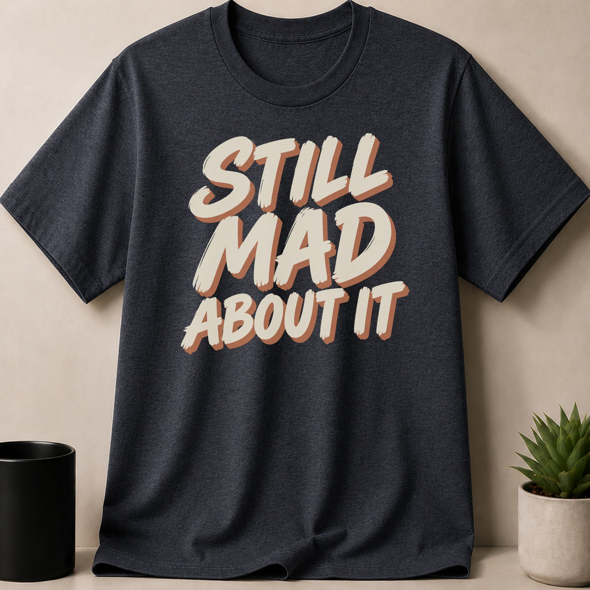 Dark grey t-shirt with "STILL MAD ABOUT IT" text in cream and brown.