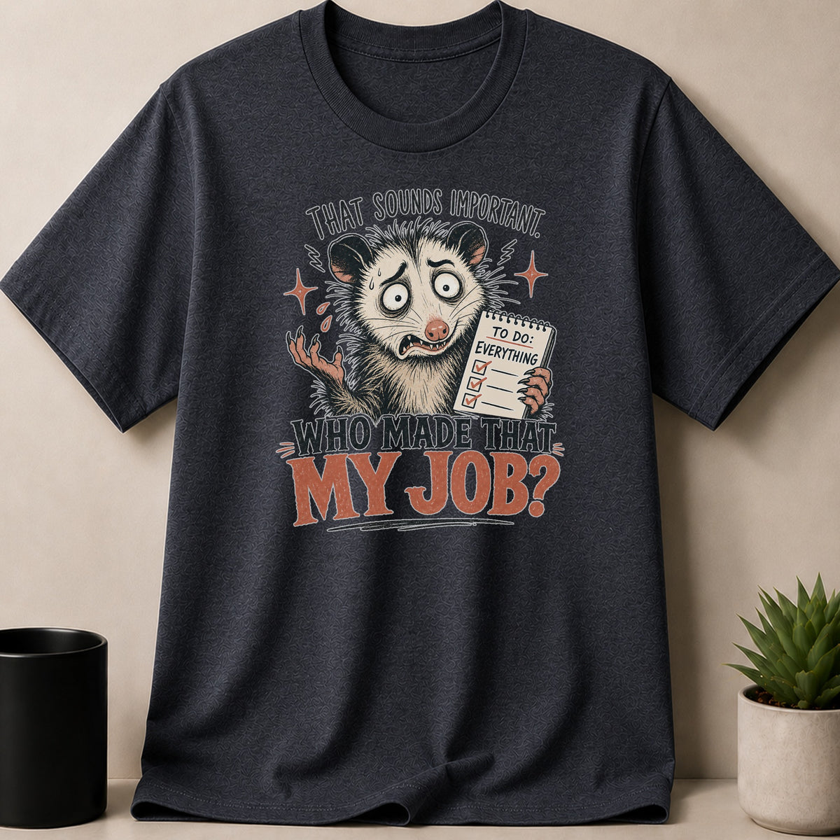 Dark grey t-shirt featuring an overwhelmed opossum with a to-do list and the text "Who made that my job?".