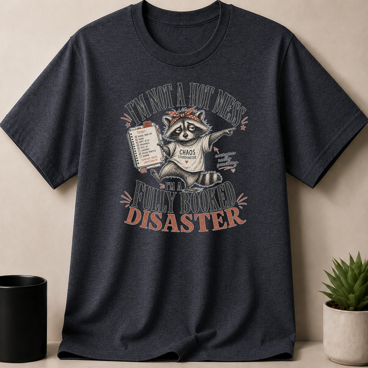 Dark Heather t-shirt — Raccoon wearing "Chaos Coordinator" shirt, holding a to-do list, "I'm Not A Hot Mess I'm a Fully Booked Disaster" text.