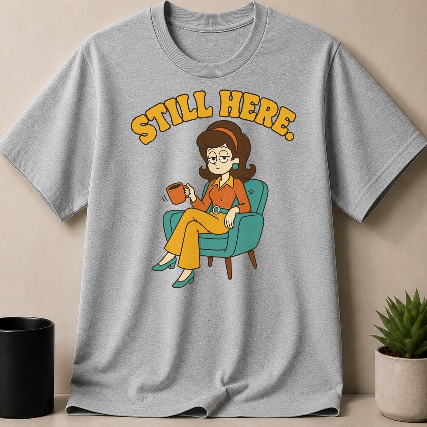 Grey t-shirt with cartoon woman in chair holding coffee and text "Still Here.".