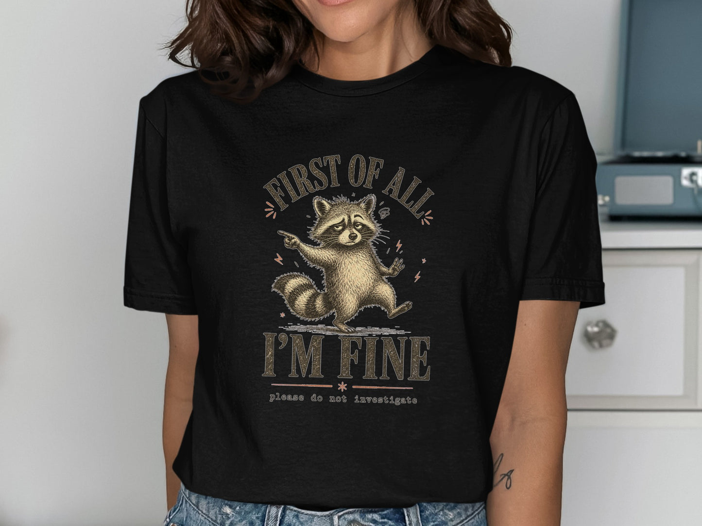 Black t-shirt with raccoon illustration and text "FIRST OF ALL I'M FINE please do not investigate".