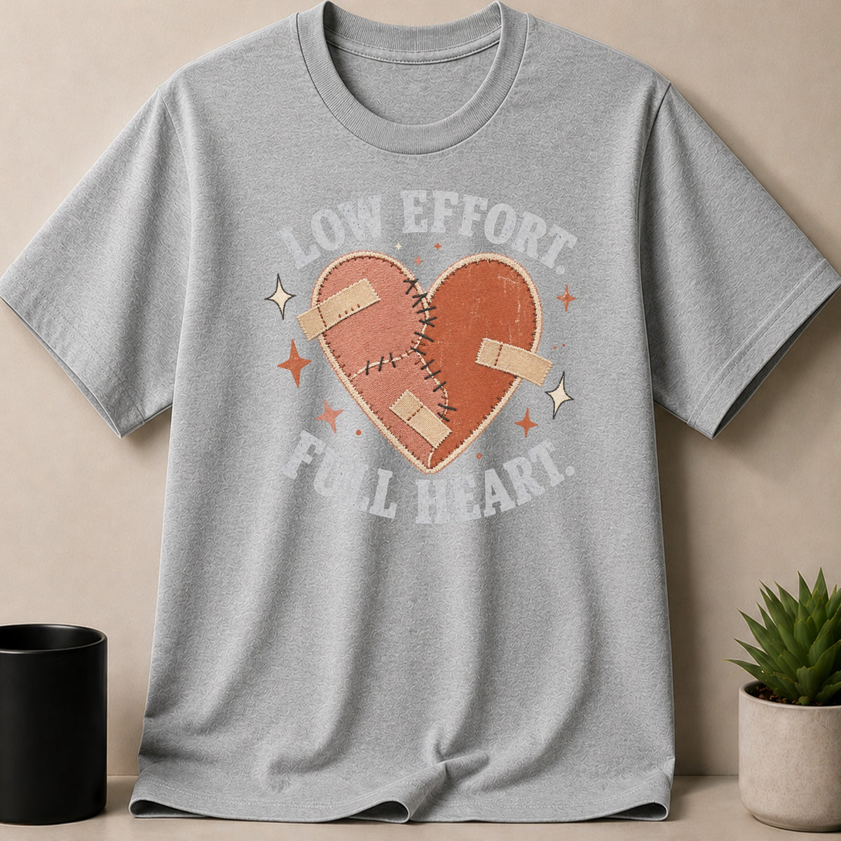Light gray t-shirt with a stitched heart, band-aids, and text "LOW EFFORT FULL HEART."