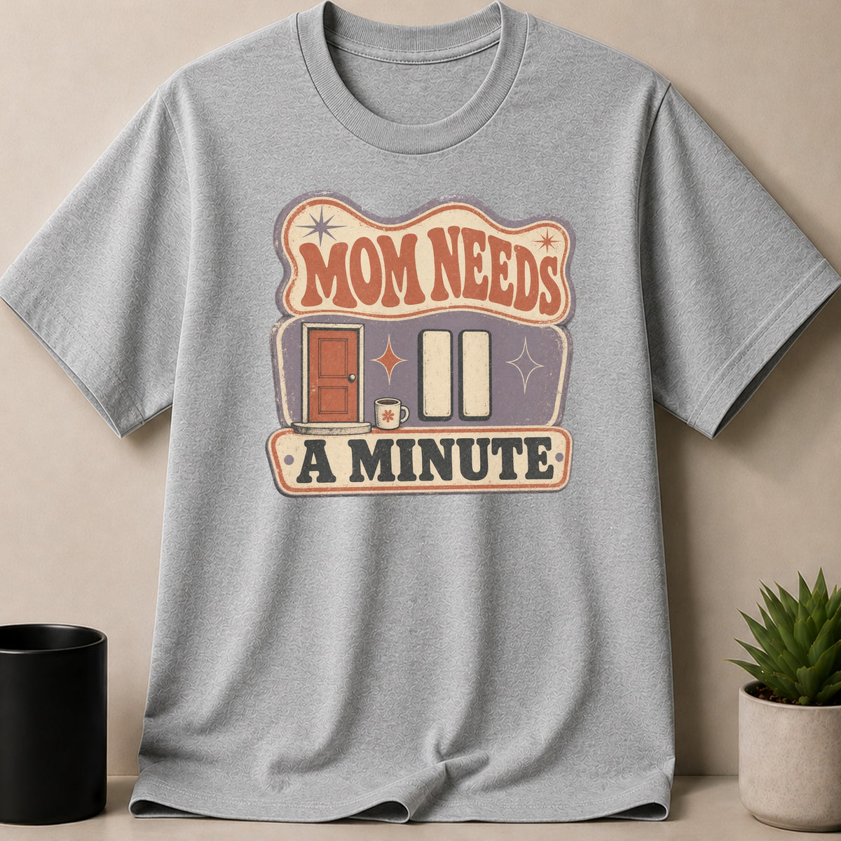 Grey t-shirt with "MOM NEEDS A MINUTE" graphic featuring a door, coffee cup, and pause buttons.