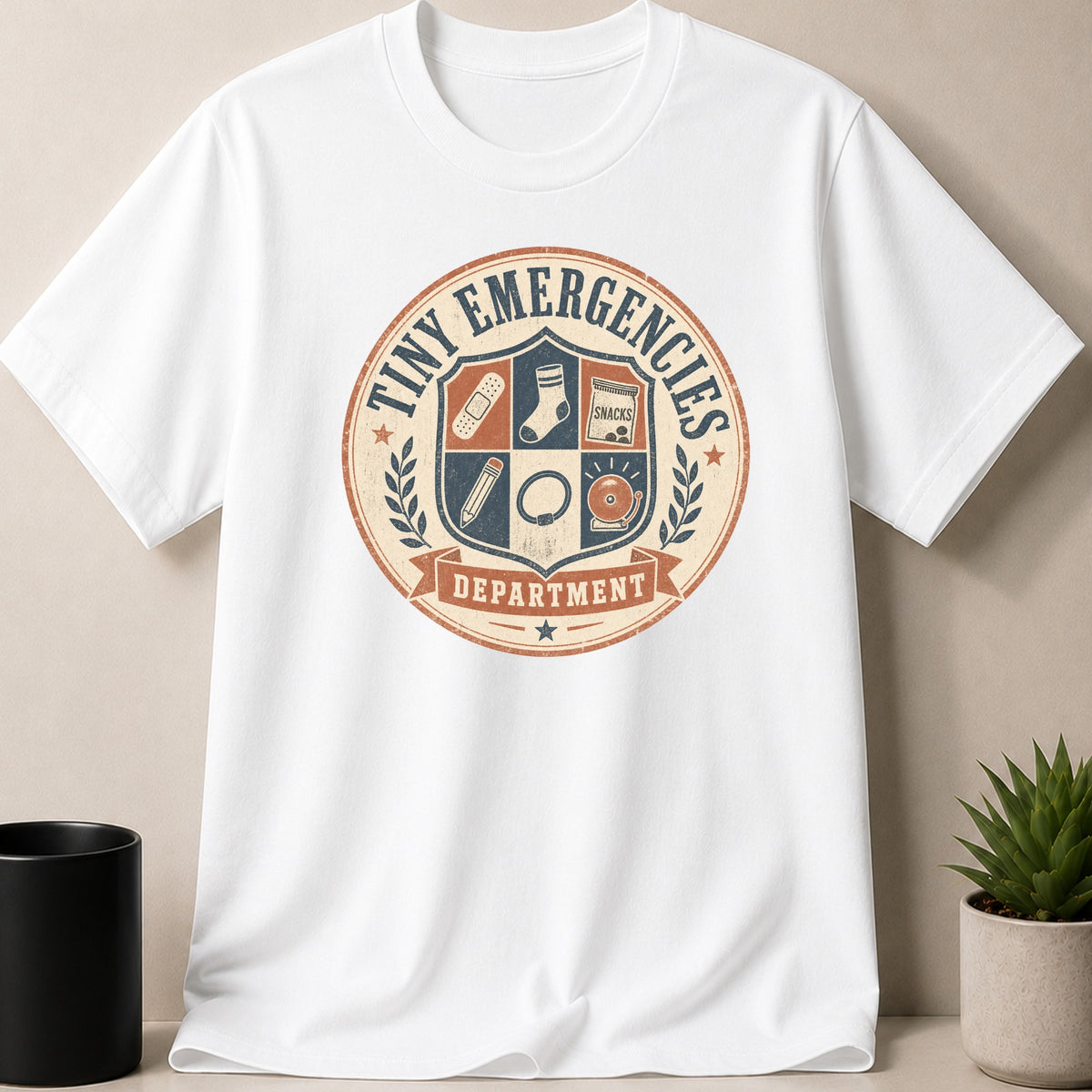 White t-shirt with "Tiny Emergencies Department" crest graphic featuring band-aids, sock, and snacks.