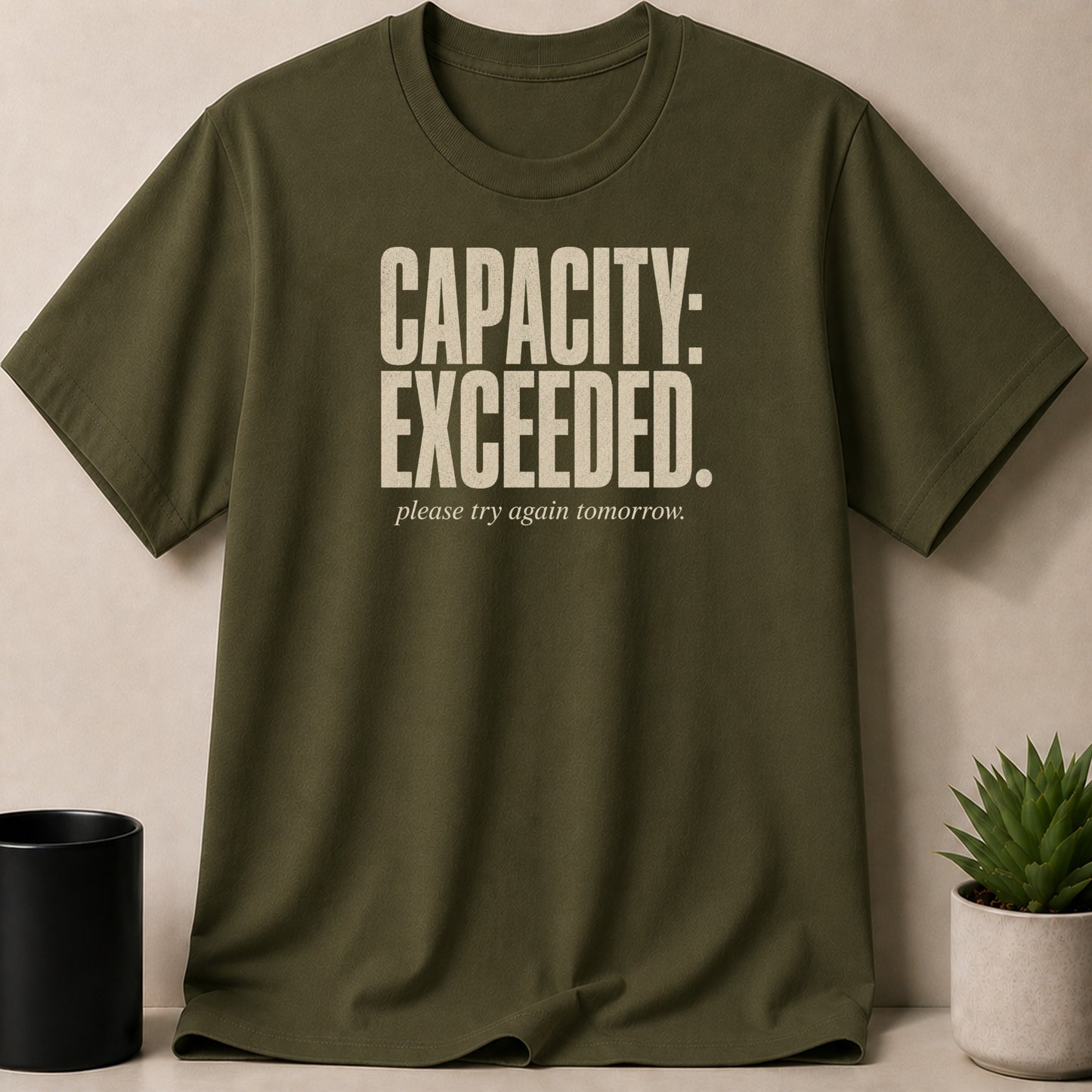 Olive green t-shirt with cream text reading "CAPACITY: EXCEEDED. please try again tomorrow."
