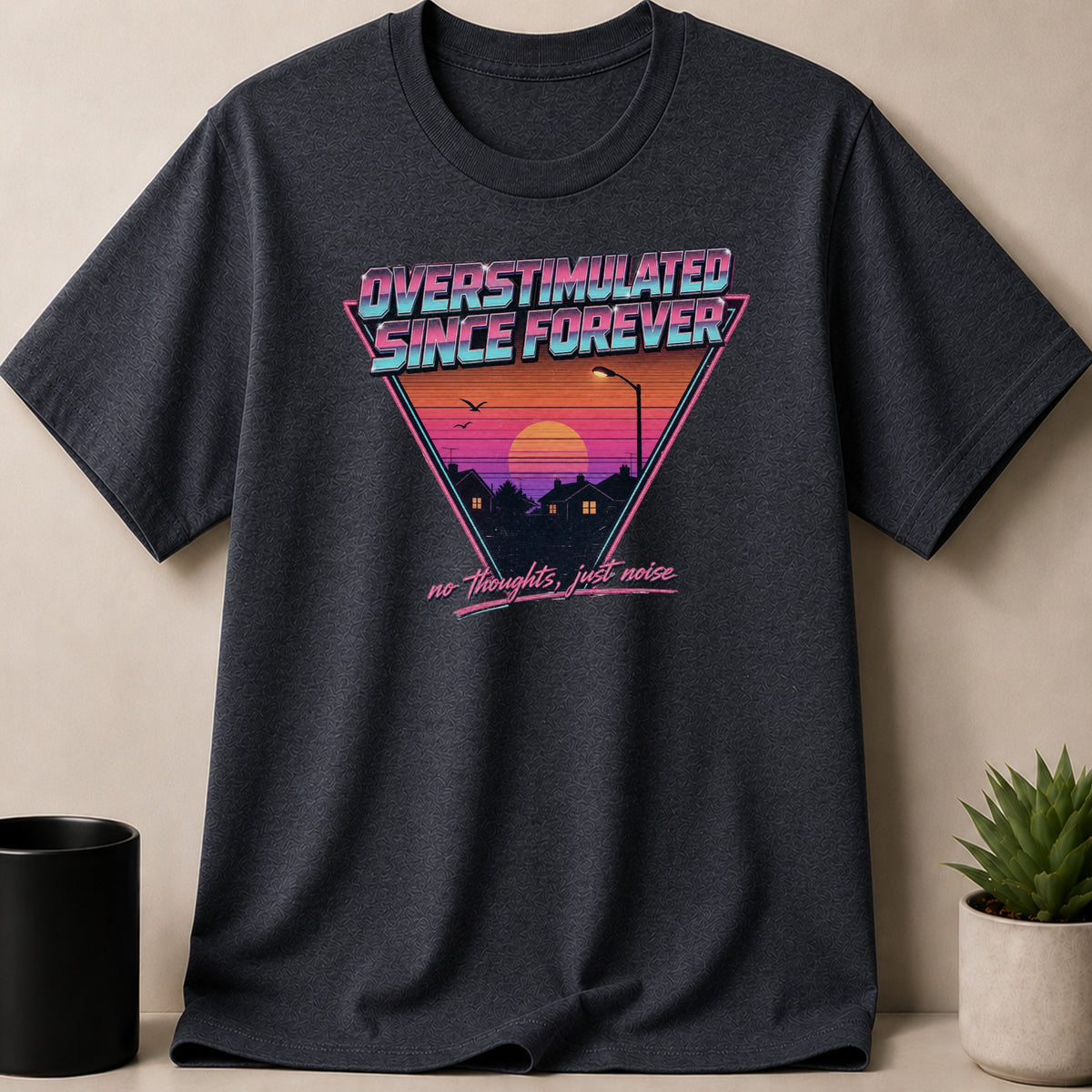 Dark t-shirt with triangle graphic: "OVERSTIMULATED SINCE FOREVER", sunset, houses and trees.
