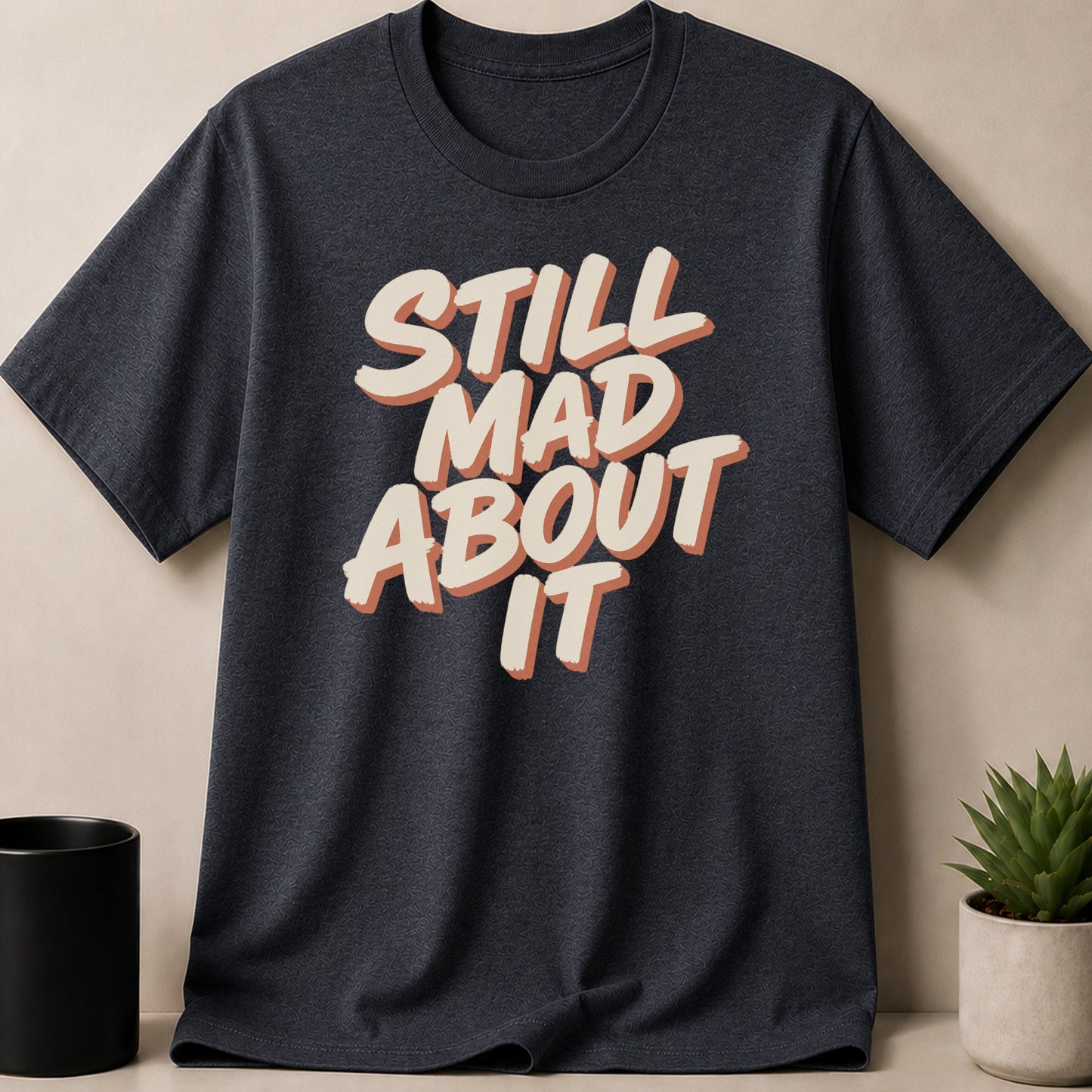 Dark t-shirt with cream and orange text reading "STILL MAD ABOUT IT".