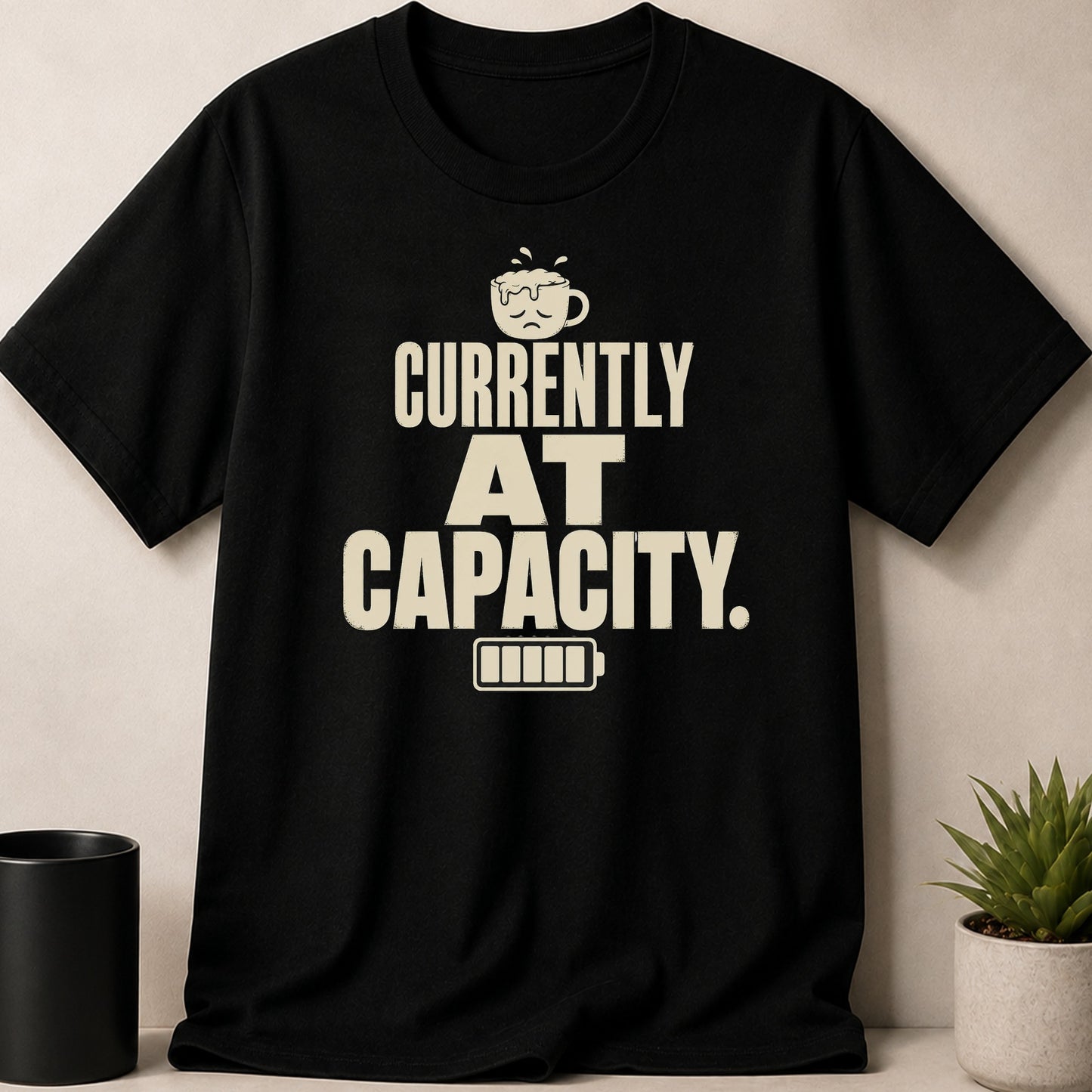 Black t-shirt with "CURRENTLY AT CAPACITY." text and a sad coffee cup icon.