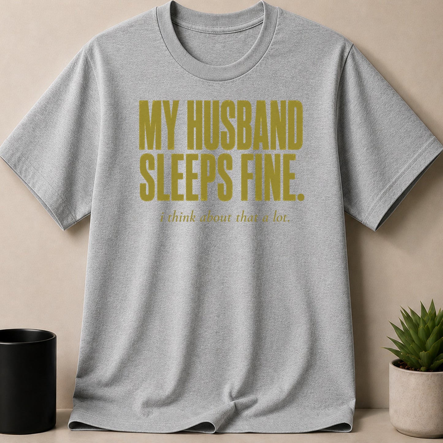 Grey t-shirt with gold text reading "MY HUSBAND SLEEPS FINE. i think about that a lot."