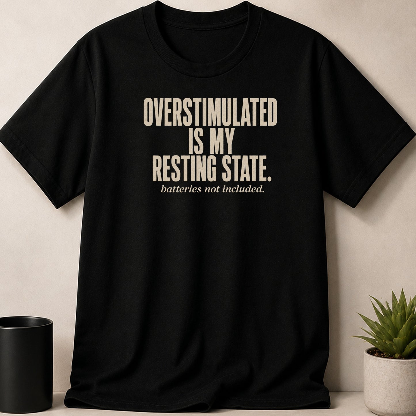 Black t-shirt with text "OVERSTIMULATED IS MY RESTING STATE. batteries not included."