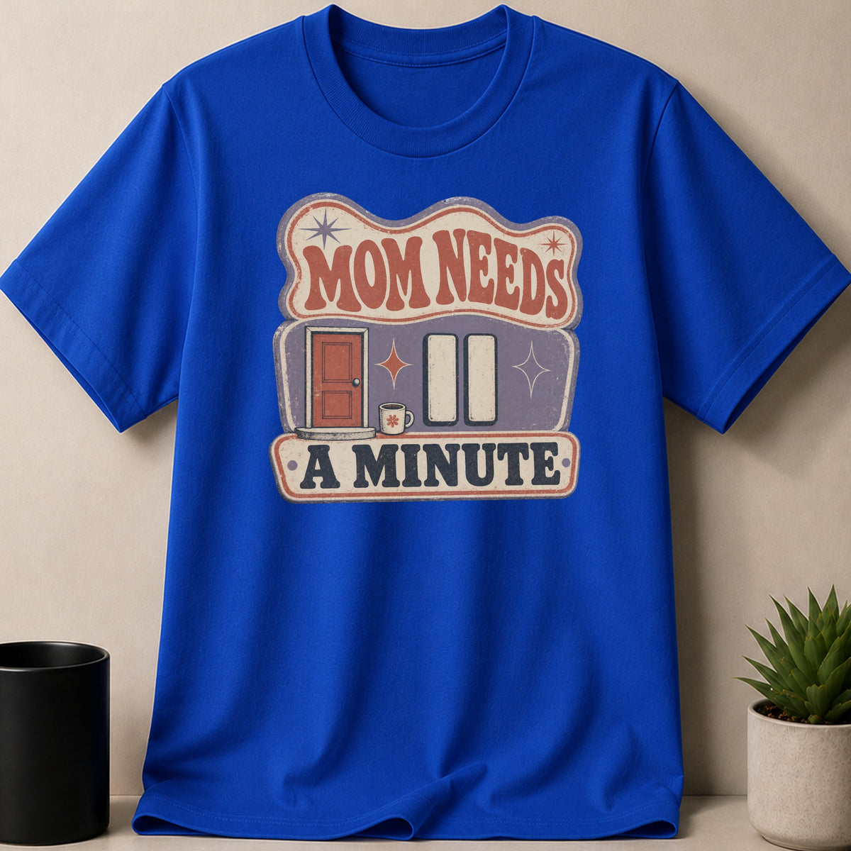 Blue t-shirt with "Mom Needs a Minute" graphic design featuring a door, coffee cup, and pause buttons.