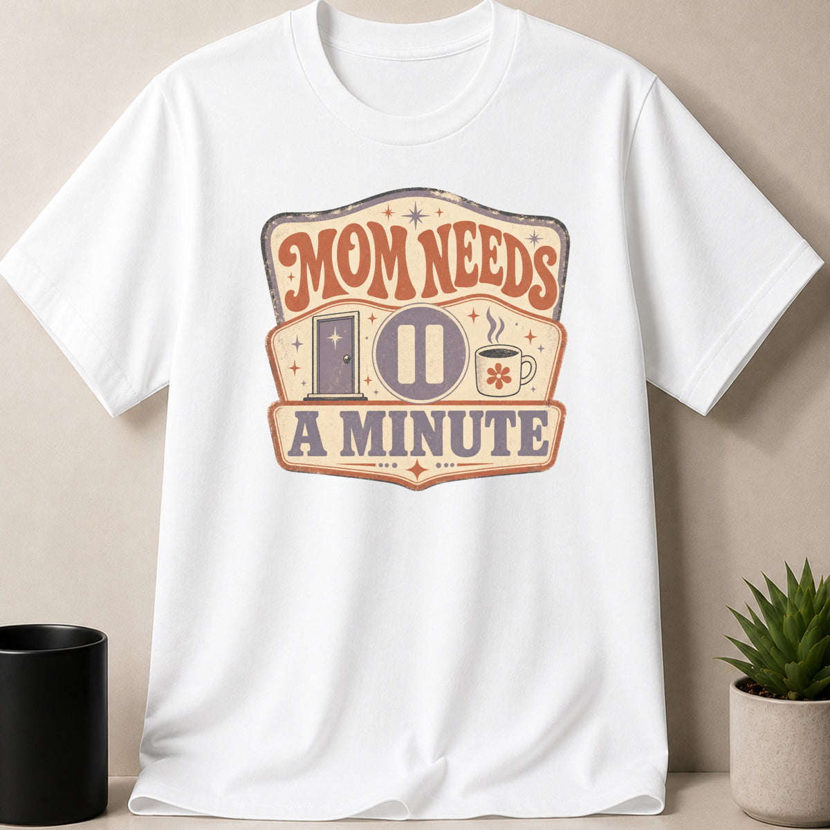 White t-shirt with "Mom Needs a Minute" graphic featuring a door, pause icon, and coffee cup.