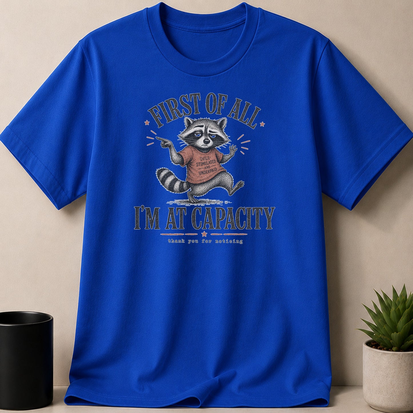 Blue t-shirt with a grumpy raccoon graphic reading "FIRST OF ALL... I'M AT CAPACITY".