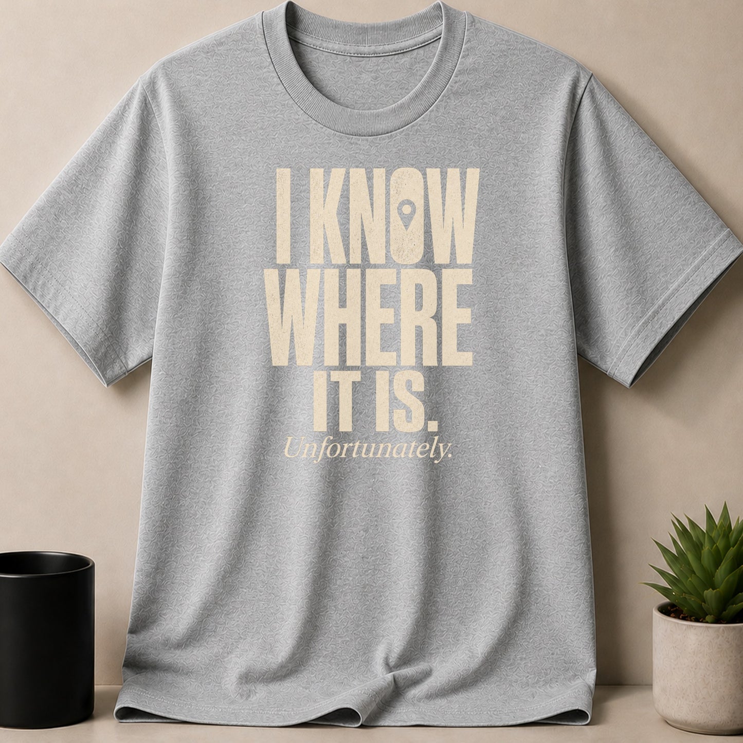 Grey t-shirt with beige text "I KNOW WHERE IT IS. Unfortunately." and a map pin icon.