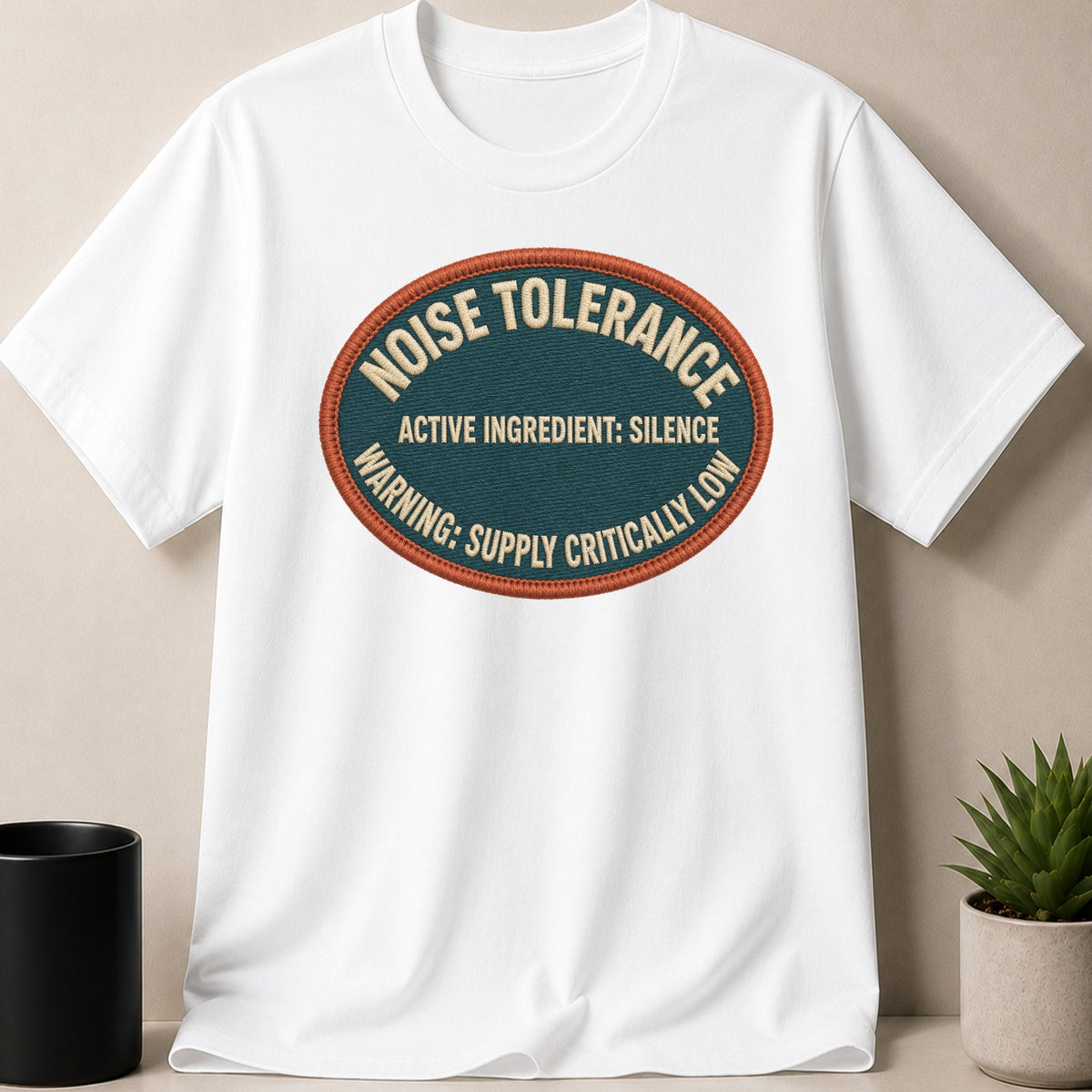 White t-shirt with oval patch reading "NOISE TOLERANCE ACTIVE INGREDIENT: SILENCE WARNING: SUPPLY CRITICALLY LOW".