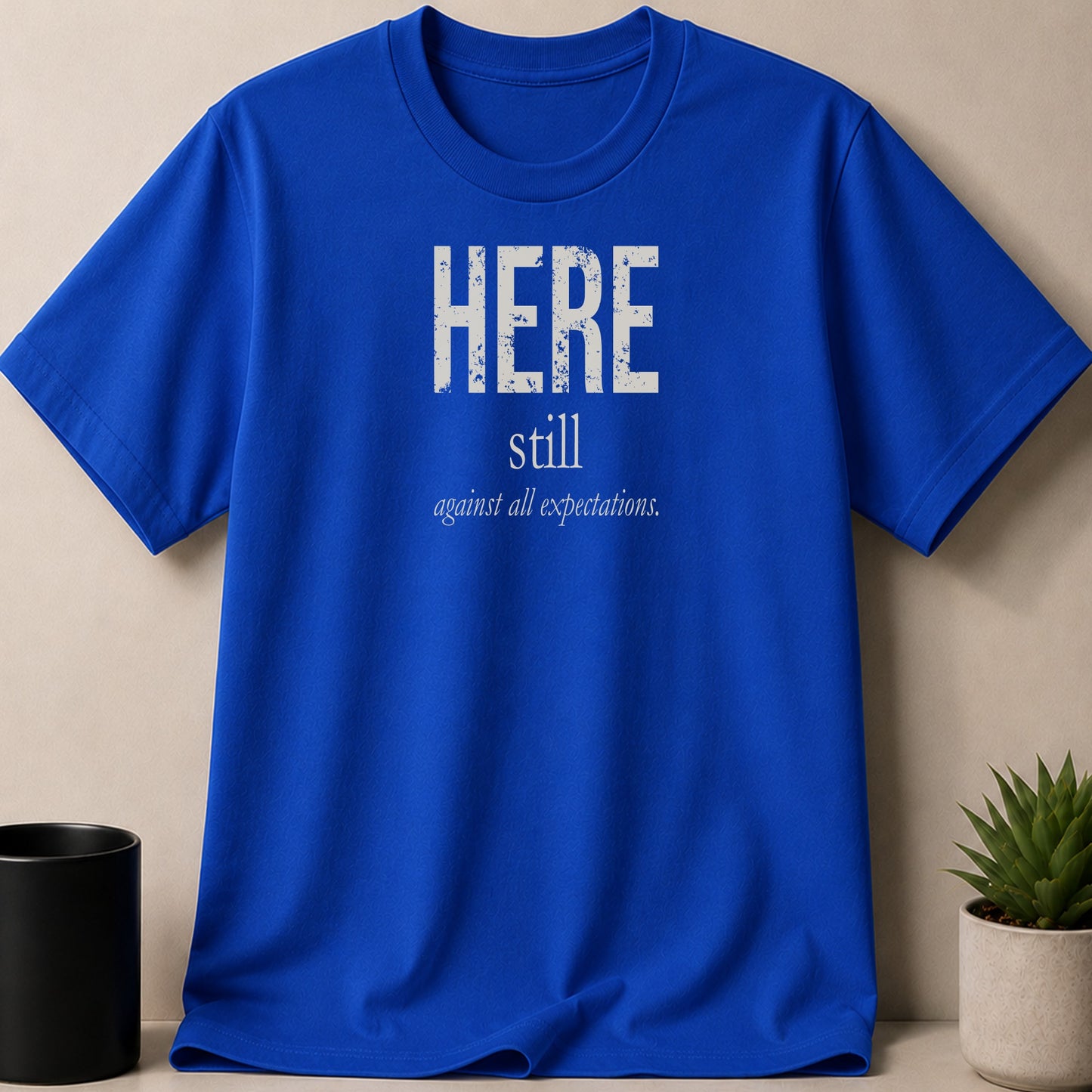 Royal blue t-shirt with white distressed text "HERE still against all expectations."