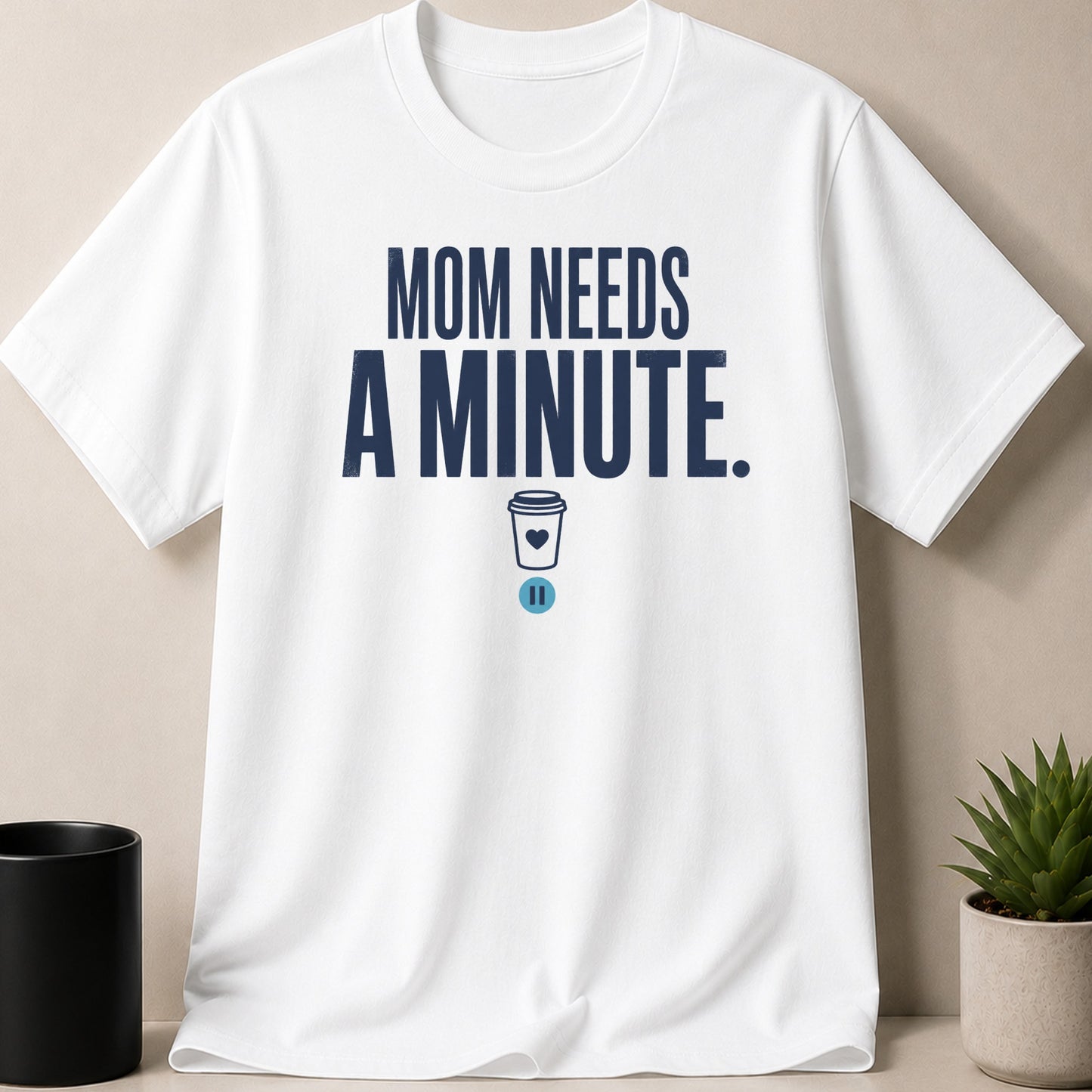 White t-shirt with dark blue text "MOM NEEDS A MINUTE." and a coffee cup icon.