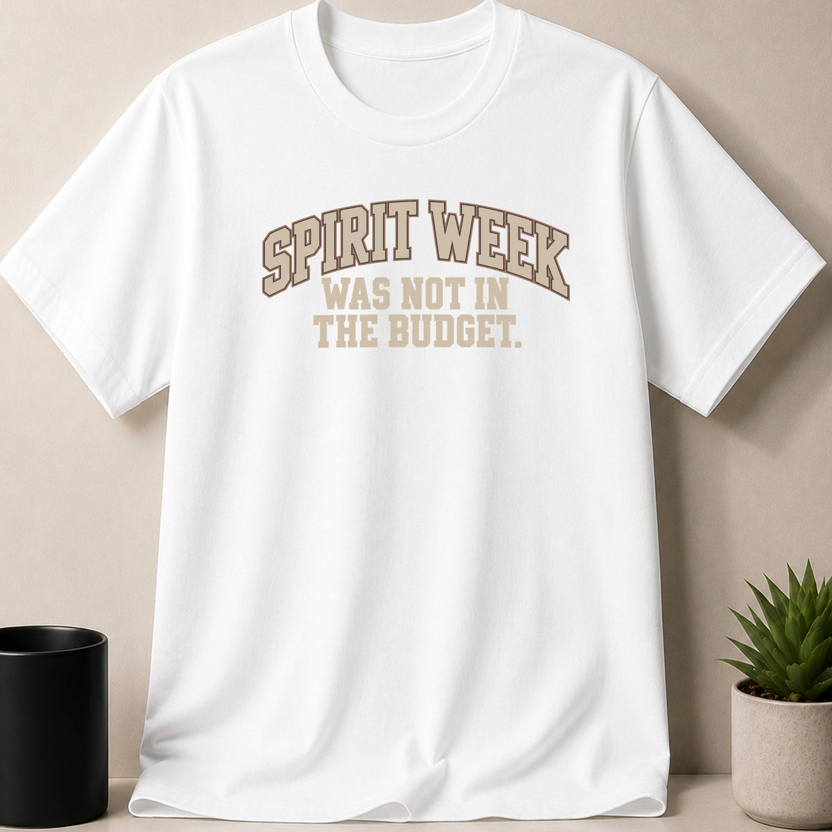 A white short-sleeved t-shirt features brown text reading SPIRIT WEEK WAS NOT IN THE BUDGET.