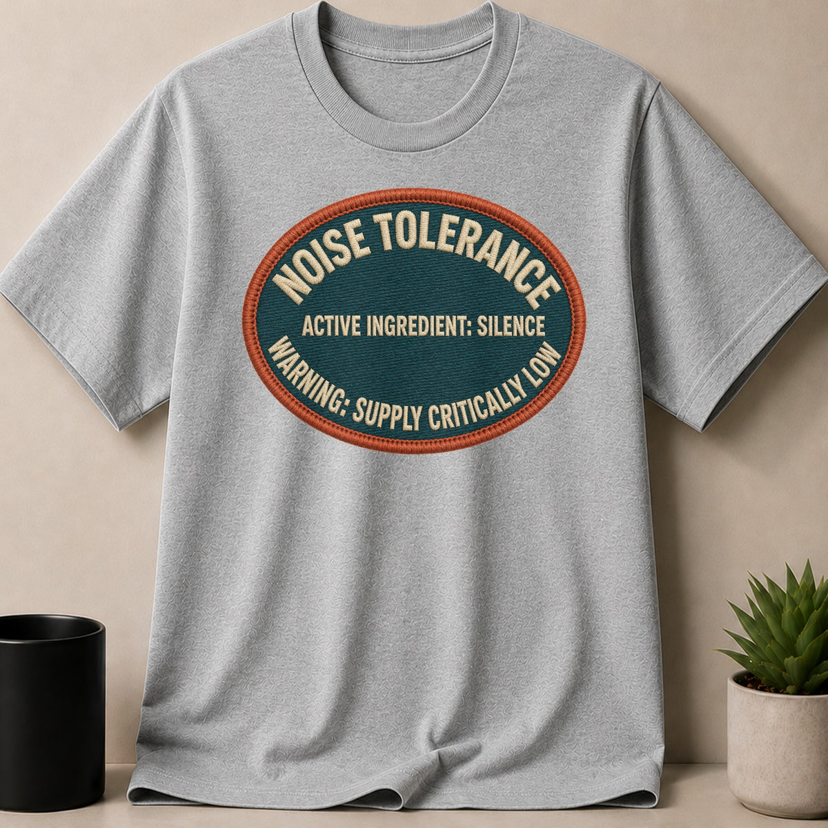 Grey t-shirt with oval patch reading "Noise Tolerance," "Active Ingredient: Silence," and "Warning: Supply Critically Low."