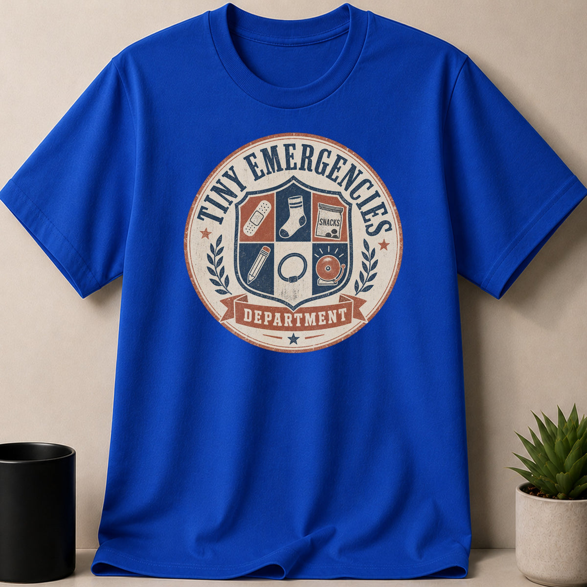 Blue t-shirt with "Tiny Emergencies Department" crest featuring band-aids, sock, snacks, pencil, ring, and bell.