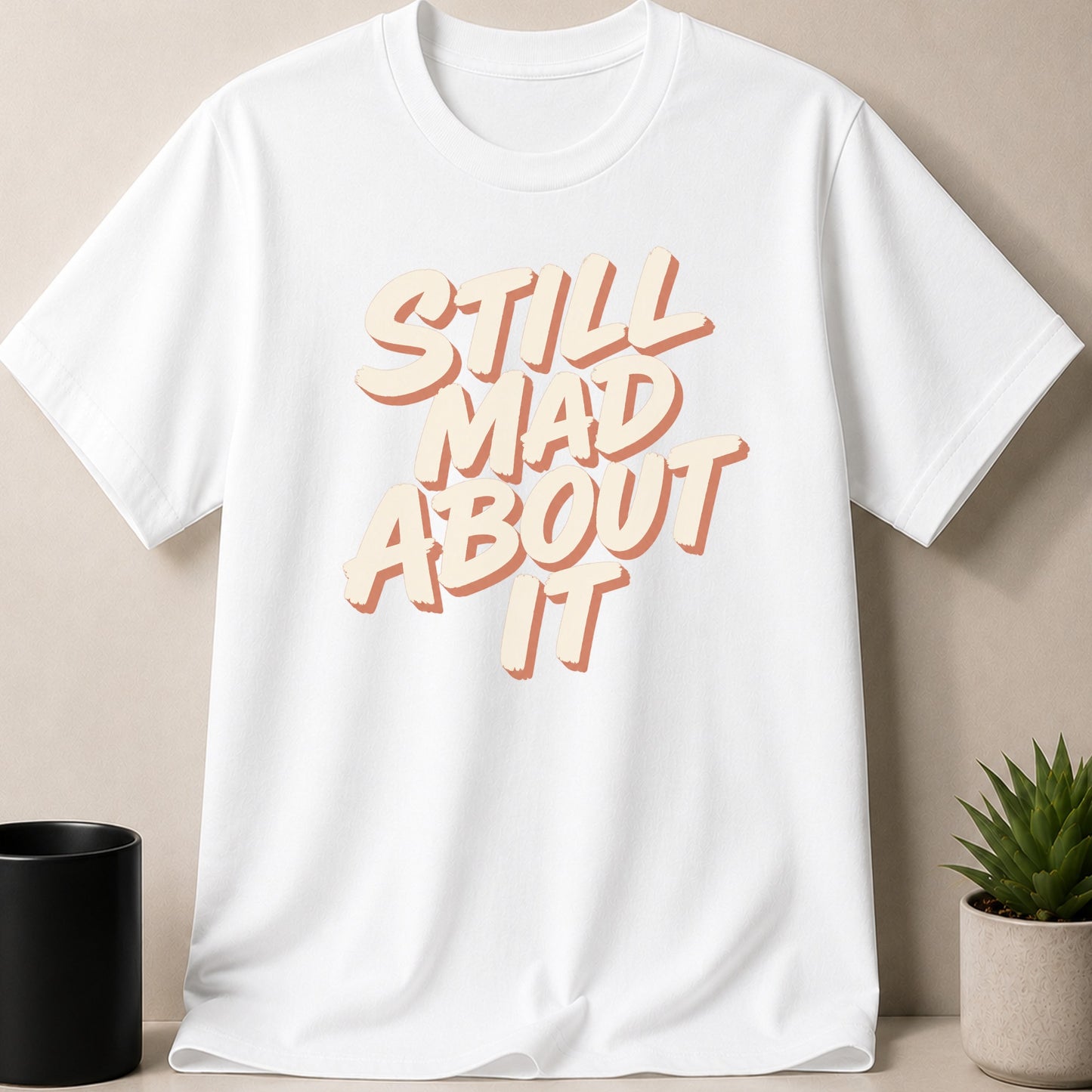 White t-shirt with "Still Mad About It" text in layered cream and orange font.