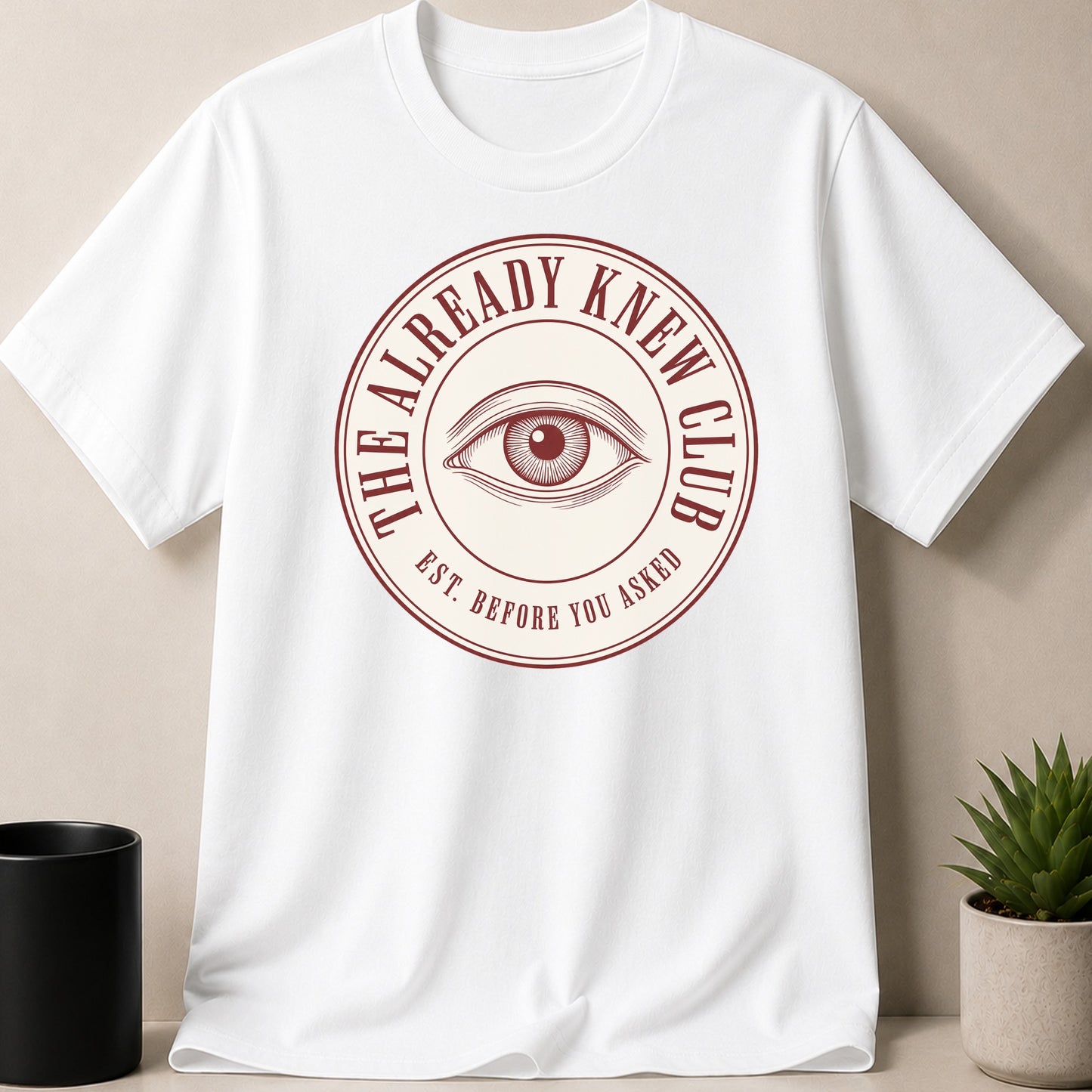 White t-shirt with a circular red graphic featuring an eye and text "The Already Knew Club Est. Before You Asked".