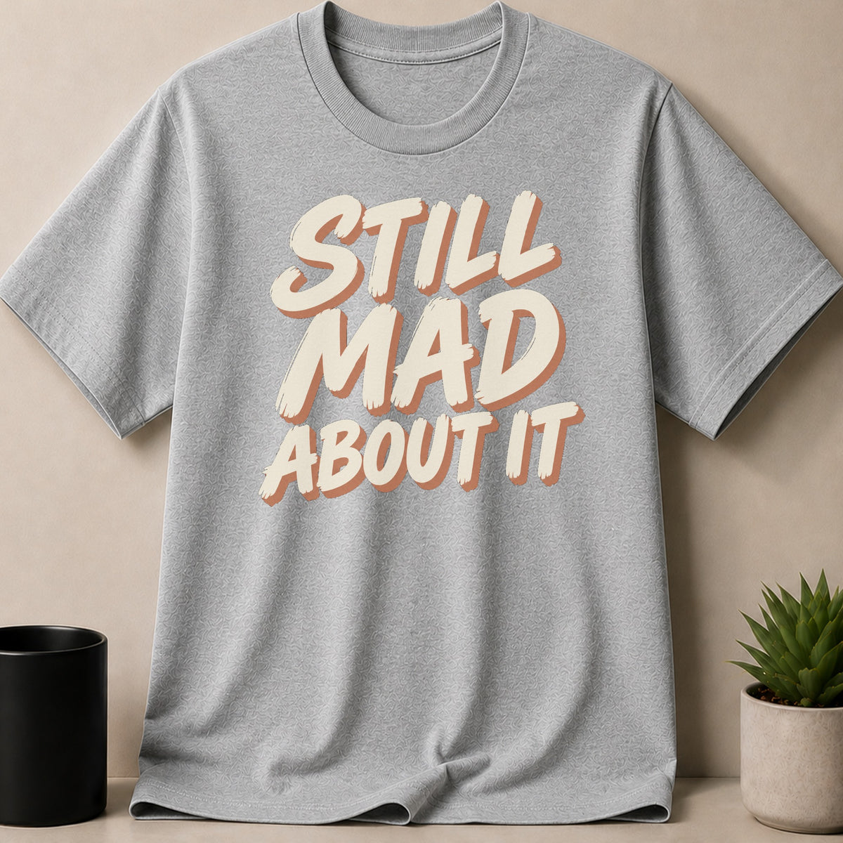 Grey t-shirt with the text "STILL MAD ABOUT IT" in layered lettering.