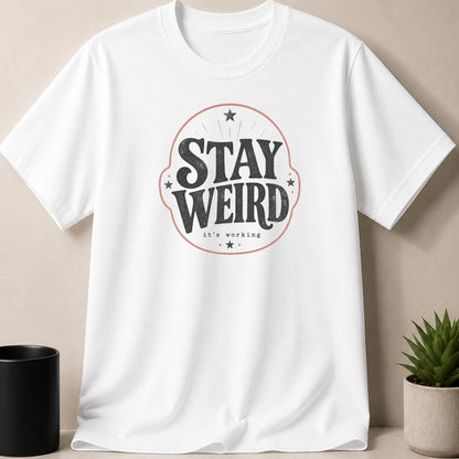 White t-shirt with "STAY WEIRD it's working" text and decorative elements.