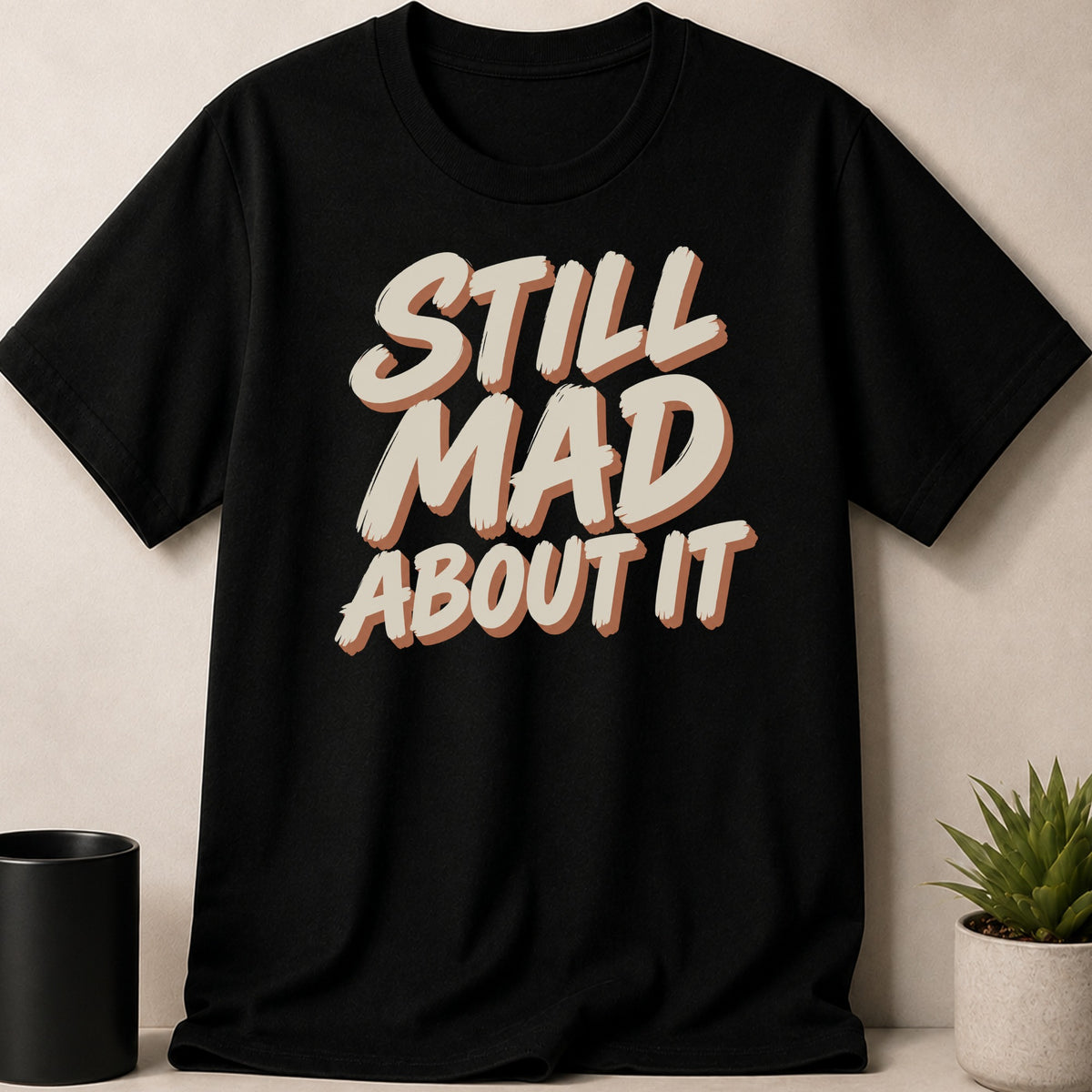 Black t-shirt with "STILL MAD ABOUT IT" text in beige and brown.