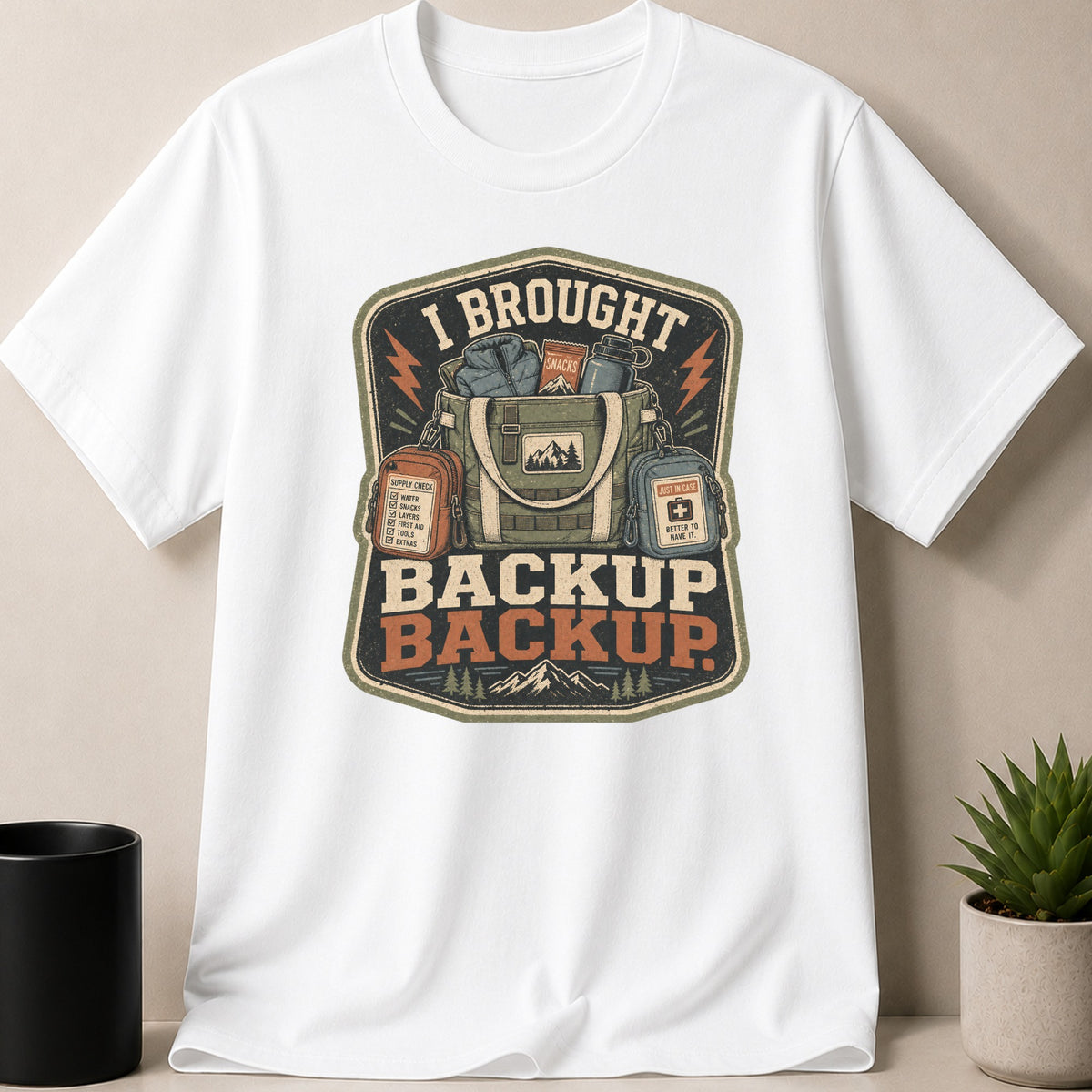 White t-shirt with "I BROUGHT BACKUP BACKUP" graphic featuring outdoor gear and mountains.