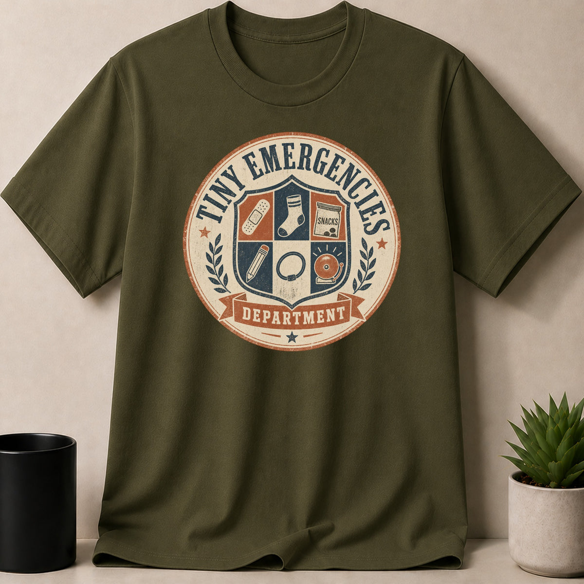 Green t-shirt with "Tiny Emergencies Department" crest featuring band-aid, sock, snacks, pencil, ring, and bell.