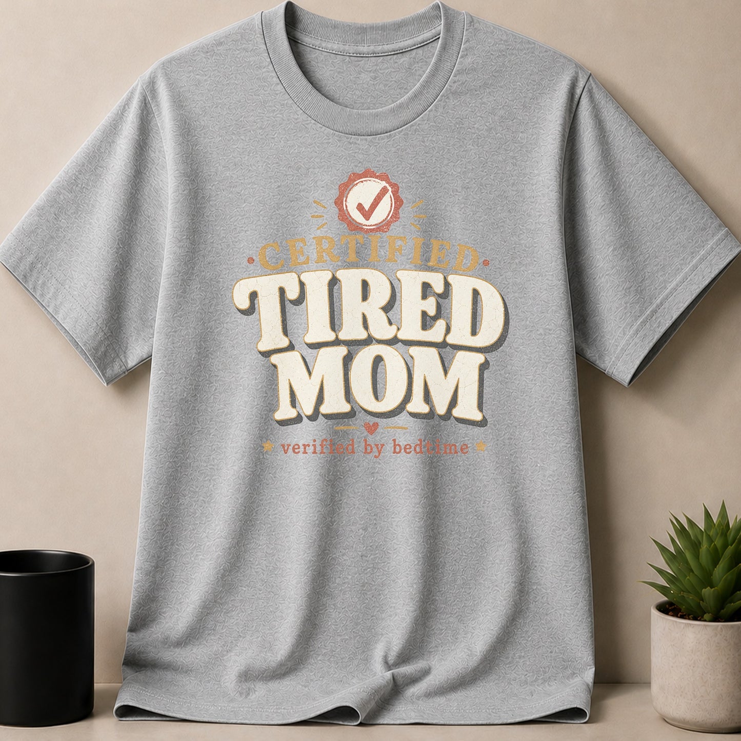 Gray t-shirt with "Certified Tired Mom verified by bedtime" text and a checkmark emblem.