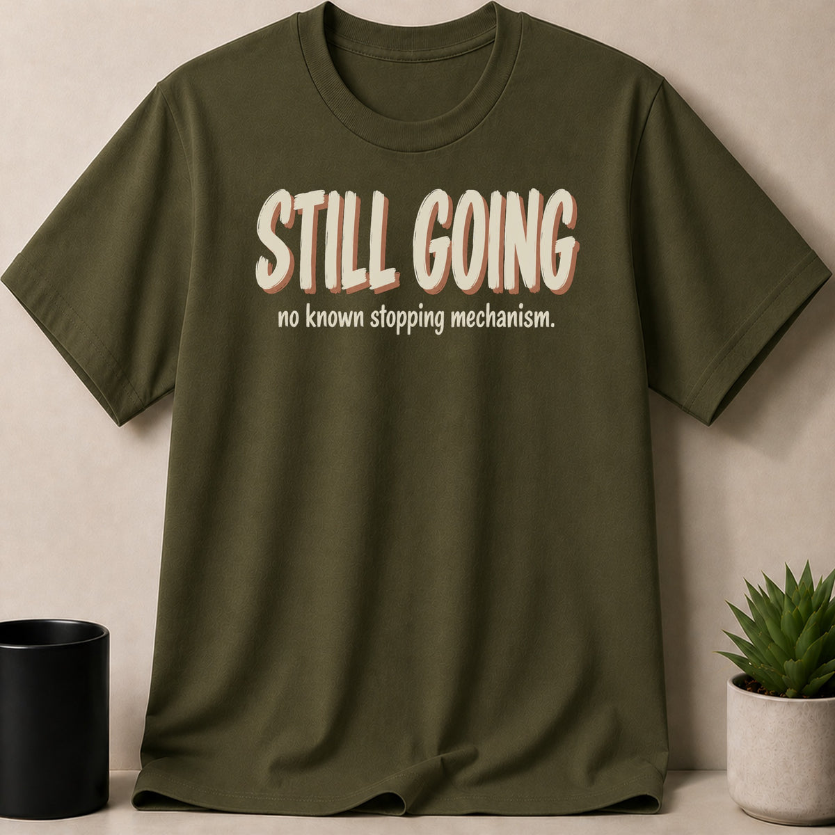 Olive green t-shirt with "STILL GOING" and "no known stopping mechanism" text.