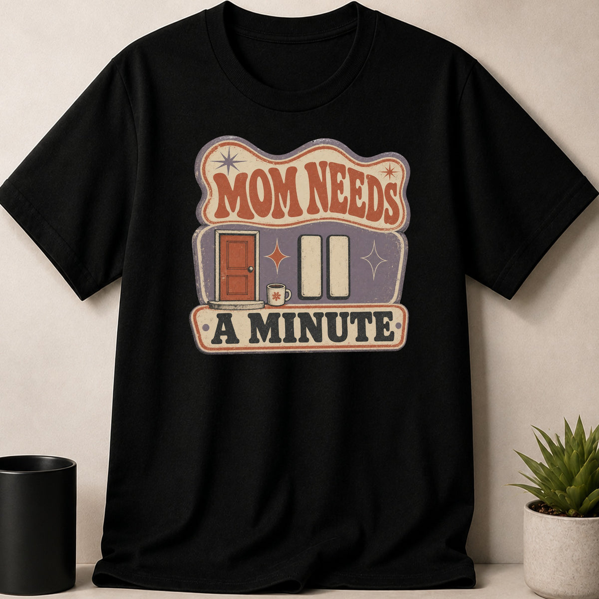 Black t-shirt with "MOM NEEDS A MINUTE" text and graphic design.