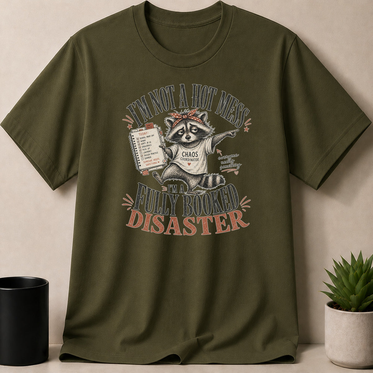 Military Green t-shirt — T-shirt with a raccoon wearing a bandana and a "Chaos Coordinator" shirt. Text reads "I'm not a hot mess, I'm a fully booked disaster."