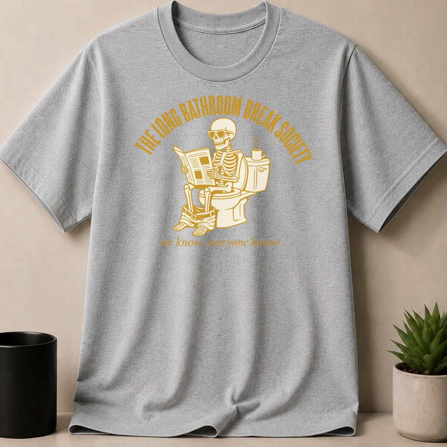Grey t-shirt with yellow graphic of skeleton on toilet reading newspaper.