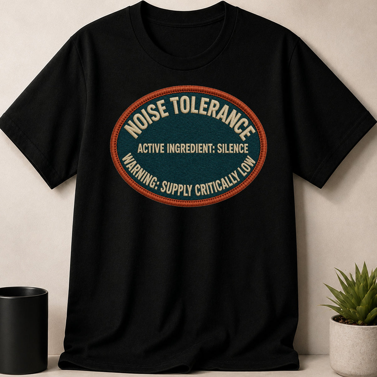 Black t-shirt with oval patch reading: Noise Tolerance, Active Ingredient: Silence, Warning: Supply Critically Low.