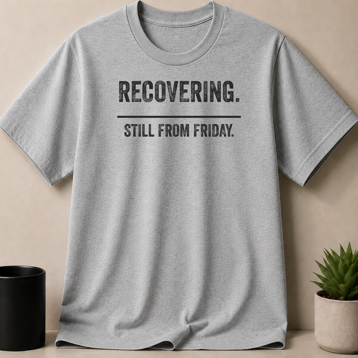 A grey t-shirt featuring the text RECOVERING. STILL FROM FRIDAY in black block letters.