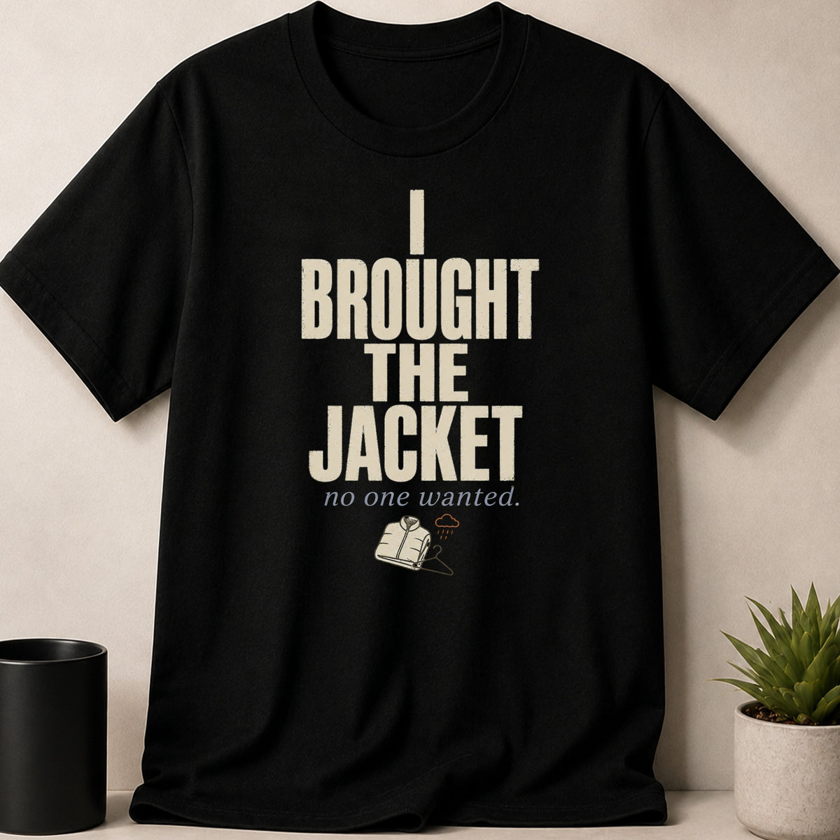Black t-shirt with cream text stating "I BROUGHT THE JACKET no one wanted." and an illustration of a jacket on a hanger with rain clouds.