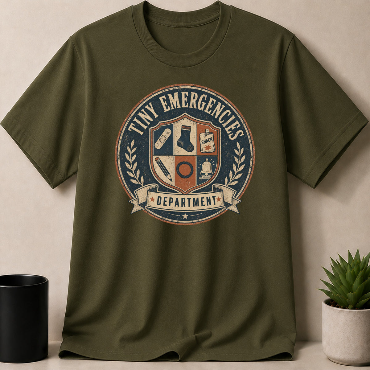 Olive t-shirt with "Tiny Emergencies Department" crest featuring a band-aid, sock, snack, pencil, and bell.