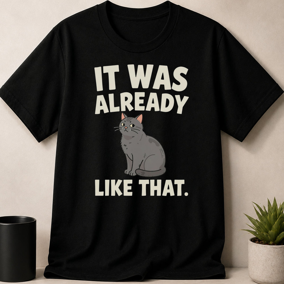 A black t-shirt printed with white text reading IT WAS ALREADY LIKE THAT and an illustration of a gray cat.