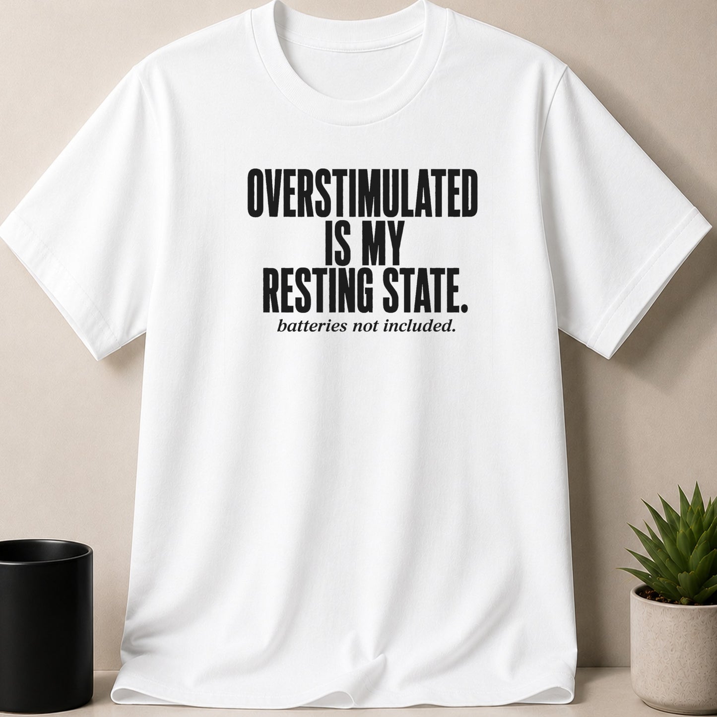 White t-shirt with black text reading "OVERSTIMULATED IS MY RESTING STATE. batteries not included."