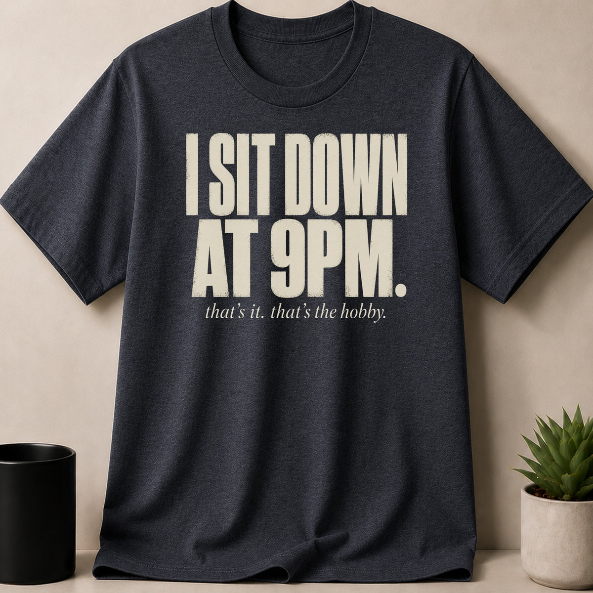 Dark gray t-shirt with cream text "I SIT DOWN AT 9PM. that's it. that's the hobby."