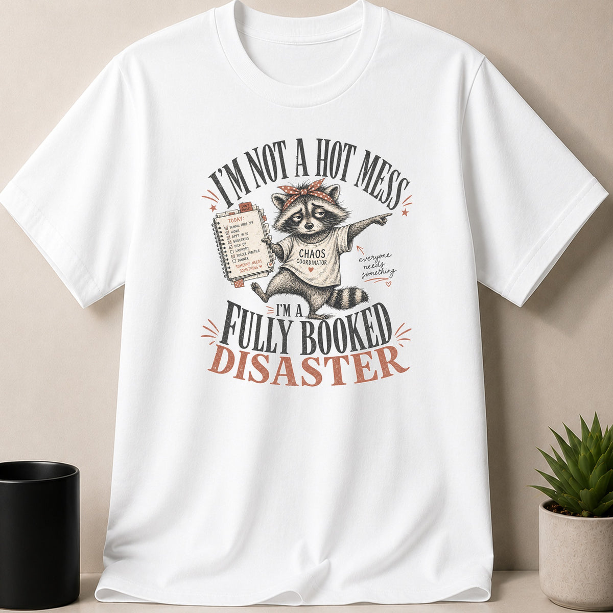 White t-shirt with raccoon graphic and text "I'm Not a Hot Mess, I'm a Fully Booked Disaster."