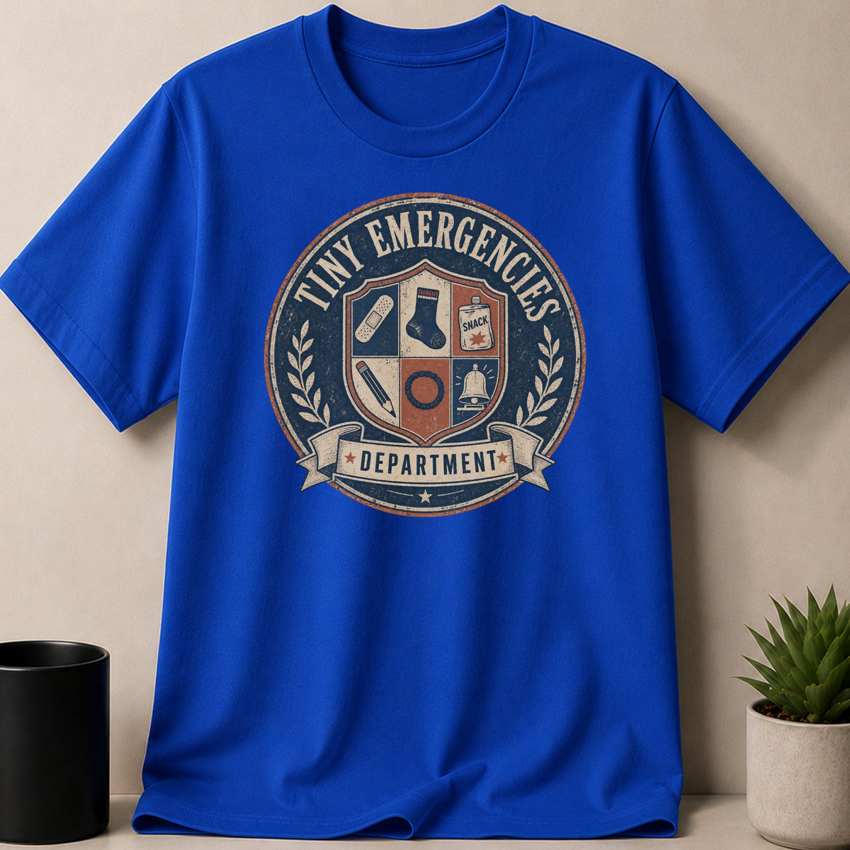 Royal blue t-shirt with "Tiny Emergencies Department" crest featuring band-aid, sock, snack, pencil, and bell.