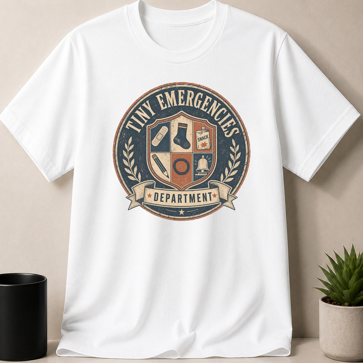 White t-shirt with "Tiny Emergencies Department" crest graphic featuring band-aid, sock, snack, pencil, ring, and bell.
