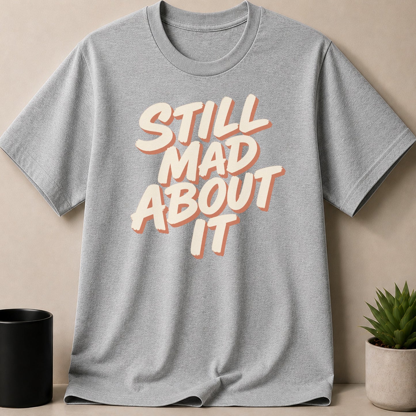 Grey t-shirt with text "STILL MAD ABOUT IT" in cream and orange.