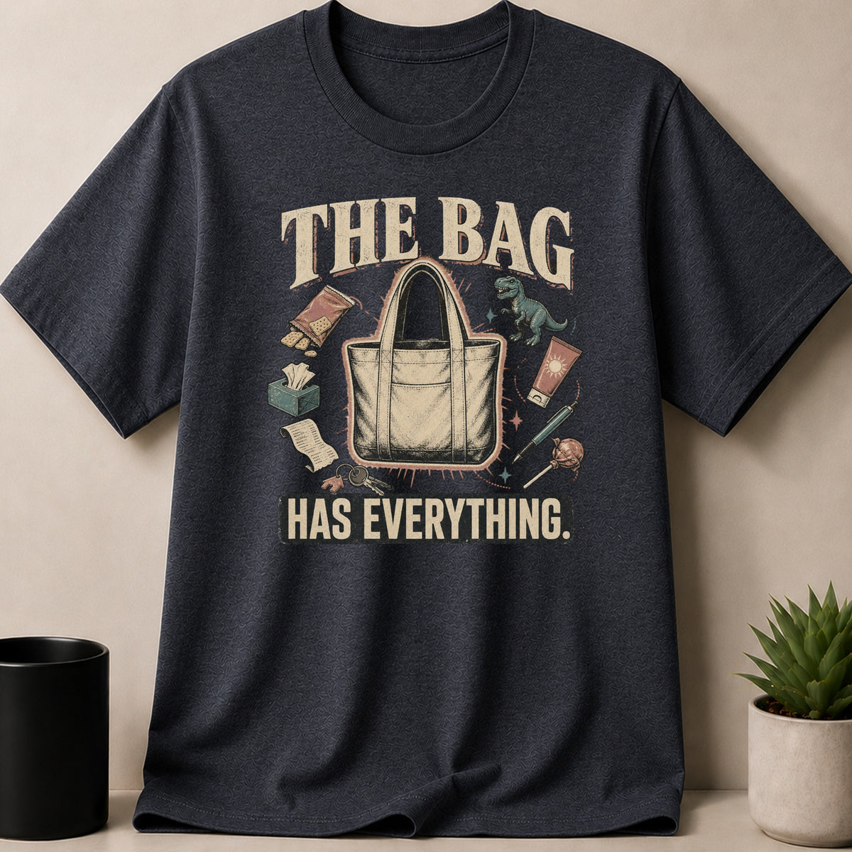 Dark Heather t-shirt — Graphic tee with "THE BAG HAS EVERYTHING" text and cartoon items around a tote bag.