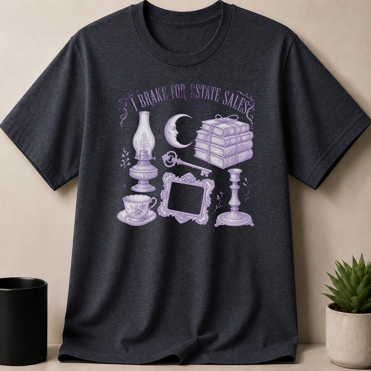 Dark t-shirt with purple illustration of oil lamp, moon, books, key, teacup, frame, and candlestick.