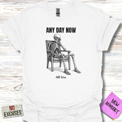 White t-shirt with skeleton sitting in chair, "ANY DAY NOW" and "still here" text.